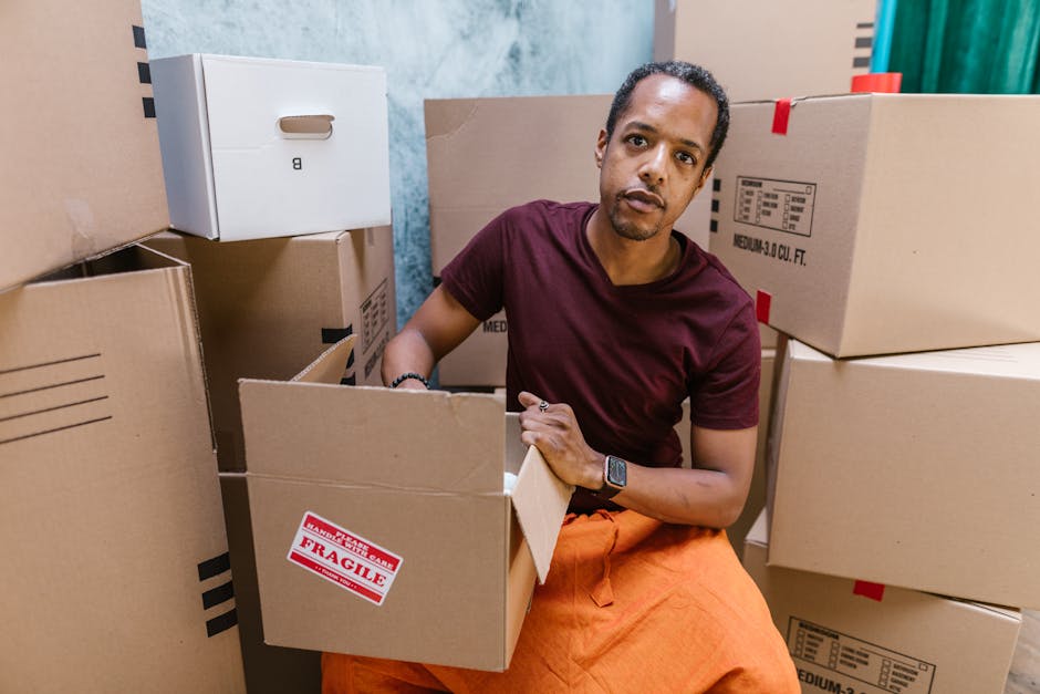 A man dressed in a maroon t-shirt and orange trousers is seated among stacked cardboard boxes inside a property. He is holding an open cardboard box with a red and white 'Fragile' sticker, indicating careful handling during a home relocation. Surrounding him are various packed boxes, some sealed with packing tape and others partially open, revealing packing materials such as bubble wrap and plastic wrapping. The boxes are positioned on the floor and some are leaning against the wall, with additional boxes stacked nearby in a cluttered manner. The environment appears to be an indoor space, possibly a living room or hallway, with partly visible walls painted in blue and other neutral tones. A moving trolley, though not directly visible, could be inferred as part of the loading process. This scene demonstrates furniture transport and packing steps involved in house removals, with a focus on careful handling and organized moving logistics that Removal Van Highbury offers for stress-free relocations.