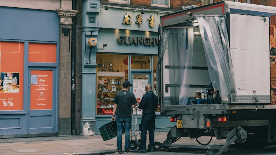 Two removal personnel from Removal Van Highbury are preparing for a home relocation outside a storefront labelled 'GUANGH' with Chinese characters above. The team is handling boxes and packaging materials, with a large moving truck parked on the pavement nearby. The truck's rear door is open, revealing a partially covered loading area with plastic sheeting to protect contents during transport. One individual is carrying a cardboard box, while the other stands nearby, possibly coordinating loading or packing tasks. The storefront has a large window displaying various items inside, and the building façade features decorative elements and signage. The scene captures a professional furniture transport and packing process, emphasizing the steps involved in residential removals, with attention to careful handling of household items before loading onto the vehicle as part of a local highbury home relocation.