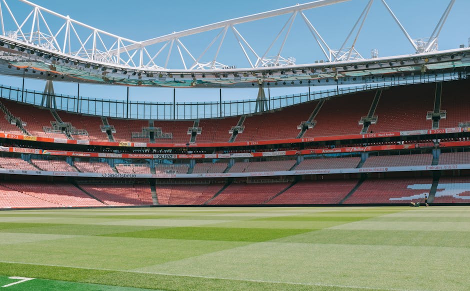 A panoramic view of a large sports stadium featuring a well-maintained grass pitch with distinct mowing patterns, surrounded by multiple tiers of empty red and pink seating areas. Above the seating, horizontal advertising banners are visible along the upper level, displaying various sponsor names and logos. The stadium's modern roof structure, composed of white steel trusses forming a geometric pattern, extends over part of the seating area, providing partial shade, supported by tall white pylons at the edges. Bright daylight illuminates the entire scene, and the sky above is clear and blue. This environment exemplifies a professional football or rugby stadium ready for match day, with no spectators present. The image provides a clear and detailed view of the stadium's architecture and layout, which can be associated with sports and large-scale events, relevant to content about venue preparation or event logistics.