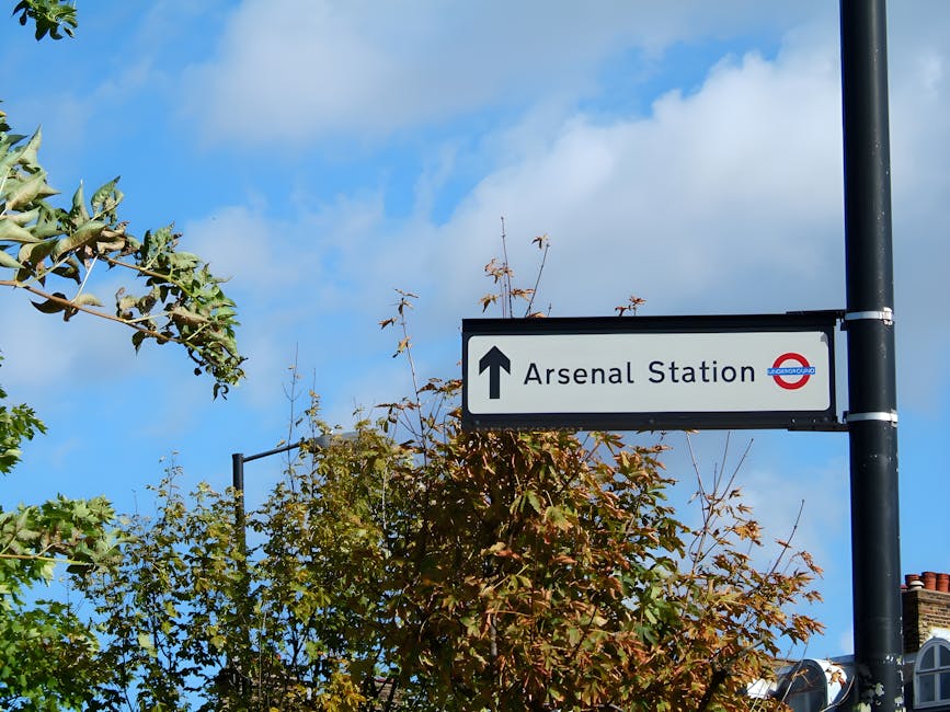 A rectangular black and white directional sign mounted on a dark metal pole points straight ahead towards Arsenal Station, a London Underground station, with the station's roundel symbol visible on the sign. The background features a blue sky with scattered white clouds and the tops of trees with green and reddish-brown leaves. In the foreground, partially visible, are the leafy branches of a tree. The scene is captured during daylight, with natural lighting highlighting the sign and surrounding foliage. This image relates to house removals and moving logistics, as signposts like this assist in navigation and planning for furniture transport and home relocation, which is relevant to the services offered by Removal Van Highbury, especially in the context of ensuring timely and efficient property moving processes.