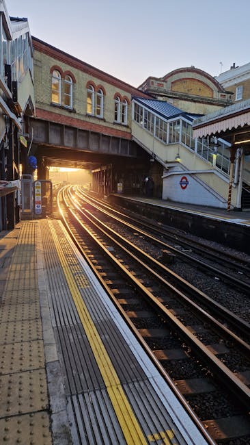 A view of a train station platform during daylight with a typical yellow tactile paving strip along the edge for safety. Metal railway tracks run parallel to the platform, leading toward a bright opening where sunlight filters through, creating a warm glow. Above the platform, there is an overpass bridge with brick and concrete construction, featuring windows and a staircase leading up to a walkway. To the right, a covered area extends with a roof supported by columns, and part of a waiting area can be seen. The scene captures the environment of a London Underground station, relevant to house removals or moving logistics involving train transportation, with no visible personnel or moving equipment.