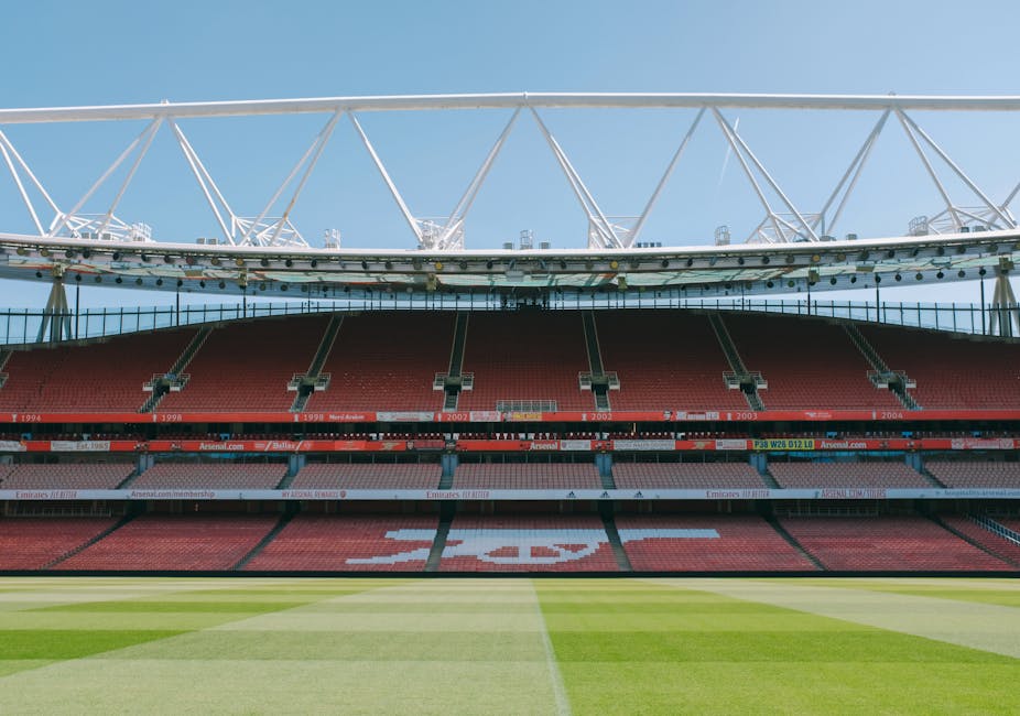 The image shows a view from inside the historic Arsenal Stadium, looking towards the empty red seating areas, with the green pitch in the foreground. The upper section features a large, modern white roof structure supported by multiple angled beams. Visible in the image is the interior of the stadium, which is well-lit with natural daylight. This setting may be relevant to home relocation or moving services, as it illustrates the iconic sports venue located near Arsenal football club. Removal Van Highbury could utilize this visual to subtly connect with house removals or furniture transport amidst such landmarks, emphasizing professional packing and loading processes in a stadium or sports arena environment.