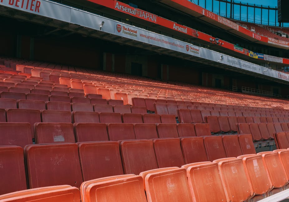 View of the empty red plastic seats in the stands of Arsenal Stadium, with the upper section featuring banners and advertisements. The seats are arranged in orderly rows and are located on an outdoor tier, illuminated by natural light. Behind the seating area, part of the stadium's exterior structure with signage referencing Arsenal Foundation and other sponsors is visible, along with a metal framework overhead. This scene captures the seating arrangement before a home relocation or moving process involving Removal Van Highbury, which provides professional removals and furniture transport services in the Highbury area, near the iconic Arsenal Stadium. The image emphasizes the stadium environment, highlighting the scale and outdoor setting typical for large-scale moving logistics related to event furniture or equipment removal.