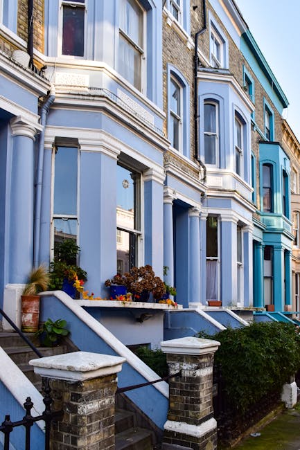 The image shows a row of Victorian-style terraced houses with ornate architectural details, painted predominantly in shades of blue and white. In front of the houses, there are brick steps leading up to the front doors, with black metal railings and small landscaped areas featuring potted plants and flowerbeds. The ground-level windows are framed with decorative molding, and some window sills have flower pots. The houses are situated on a paved sidewalk, with a clear sky overhead. This setting reflects a typical urban residential area suitable for house removals and home relocation services. The professional removals company, Removal Van Highbury, may use such a location for conducting packing, loading, and transport of household furniture and boxes during the moving process, emphasizing the importance of careful planning and efficient furniture transport within city-based house removals, especially in the Highbury area related to N5 removals advice.