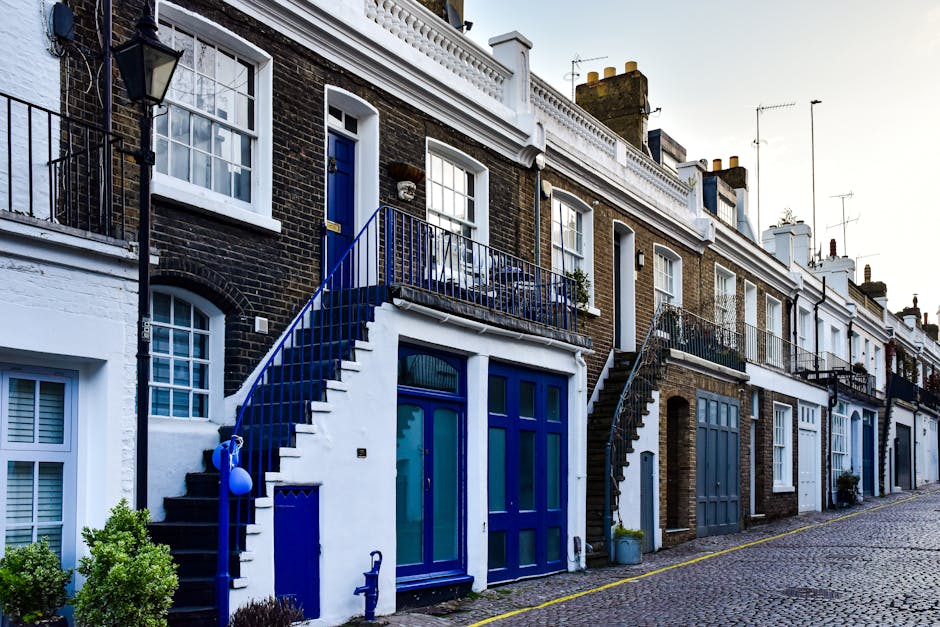 Exterior view of a row of terraced houses in Highbury with a cobblestone pavement in the foreground. The houses feature brick facades with white window frames and a mix of blue and white painted doorways. Two houses are equipped with external spiral staircases leading to small balcony areas, where furniture and potted plants are visible. A moving van from Removal Van Highbury is parked nearby, with its side door open, indicating a home relocation process. Cardboard boxes, wrapped furniture, and packing materials are being loaded onto the van, with individuals actively carrying or arranging items to facilitate furniture transport and packing during the house removals. The scene is well-lit with natural daylight, emphasizing the logistics of house removals and loading activities outside residential properties.