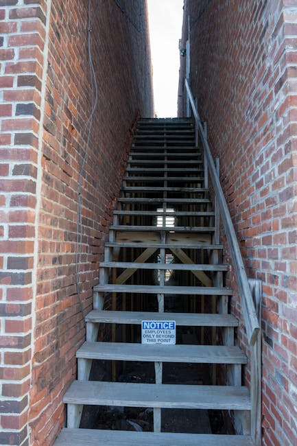 Image of an outdoor wooden staircase with narrow treads and a handrail on the right side, narrow enough to be challenging for furniture or large boxes. The staircase is situated between two tall red brick walls, leading upward to a higher level with visible gaps allowing natural light to filter through. There is a small notice sign attached to the bottom step indicating restricted access beyond that point, emphasizing the tight space involved in home relocation or moving logistics. The scene is part of a house removal process managed by Removal Van Highbury, illustrating the difficulty of navigating narrow staircases during furniture transport and packing and moving operations.