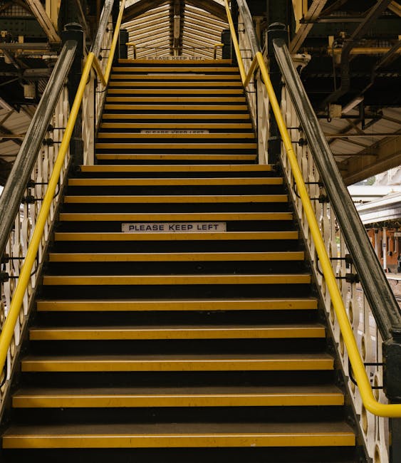 A straight staircase inside a building, leading upward with yellow handrails on each side and black treads with yellow edges. Several white stickers on the steps display the message 'PLEASE KEEP LEFT.' The staircase is illuminated by overhead lighting, revealing a metal framework and industrial elements above, suggesting a construction or industrial site environment. The setting appears used for moving or relocation activities, potentially involving furniture or packed boxes. The image captures the staircase as part of the logistics process during a home relocation, with the focus on navigating narrow stair access, an area where removals companies like Removal Van Highbury often operate to facilitate house moves within high-density residential buildings.